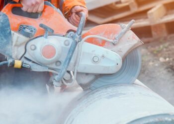 Construction worker in orange safety gear uses a handheld power saw to cut a large metal pipe, creating clouds of dust.